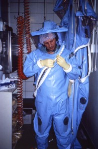  This photograph depicted a Centers for Disease Control (CDC) laboratorian, as he was putting on an older-model positive-pressure suit before entering one of the CDC’s earlier maximum containment, or Biosafety Laboratories (BSL), sometimes referred to as a “suit lab”. Scientists working within a BSL must wear protective garments that are air-tight, thereby, protecting the technician from exposure to highly-infectious pathogenic organisms. Fresh, filtered air is supplied to the interior of the suit via overhead tubing. The positive-pressurization offers some added protection to exposure through a defect in the suit, for if the suit’s patency is compromised, air would be forced out of the suit instead of being sucked into the suit.