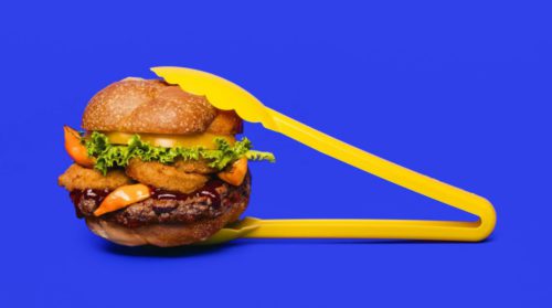 a cheese burger held by bright yellow tongs on a vivid blue background